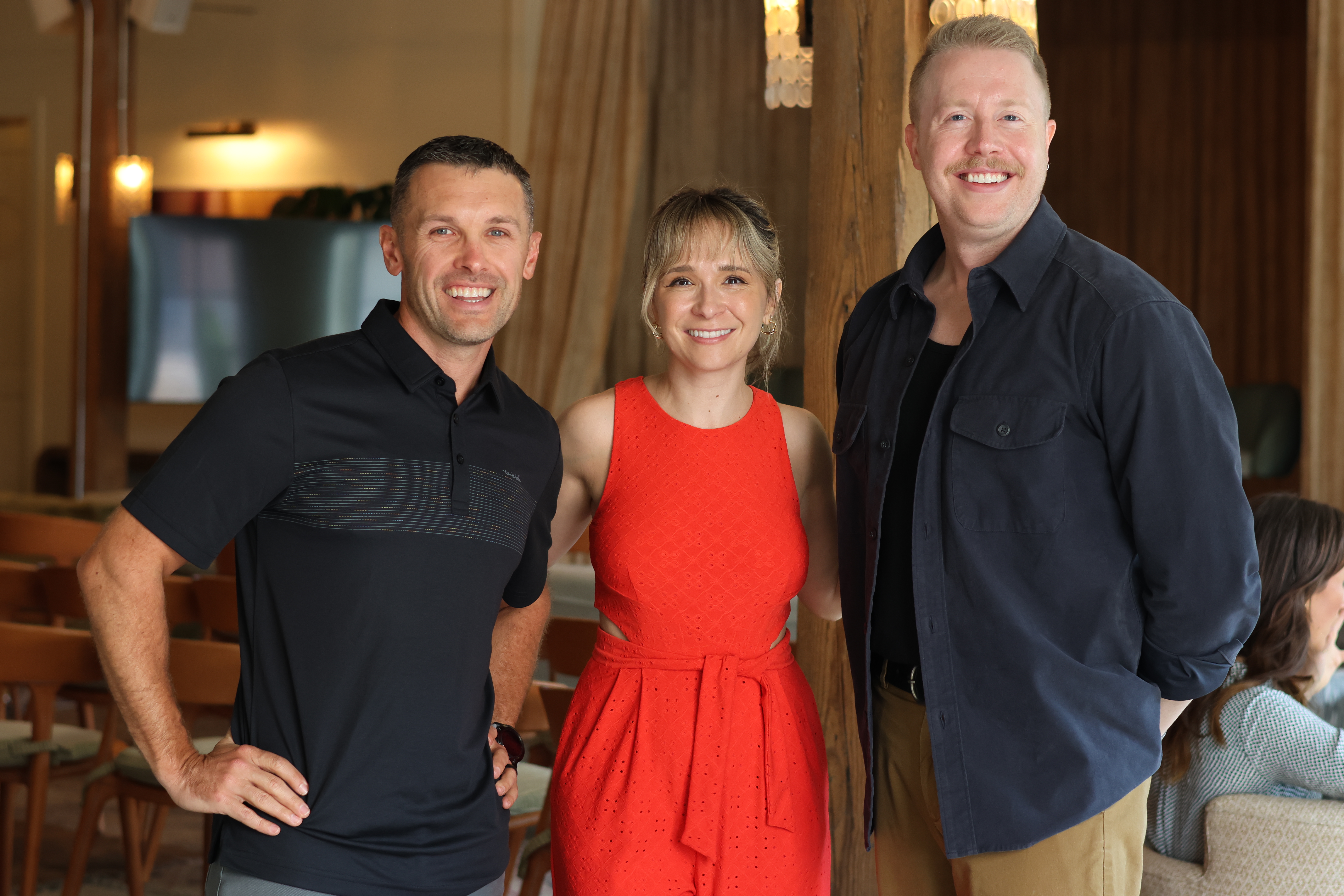 Three people stand together smiling in a well-lit, wood-paneled room.