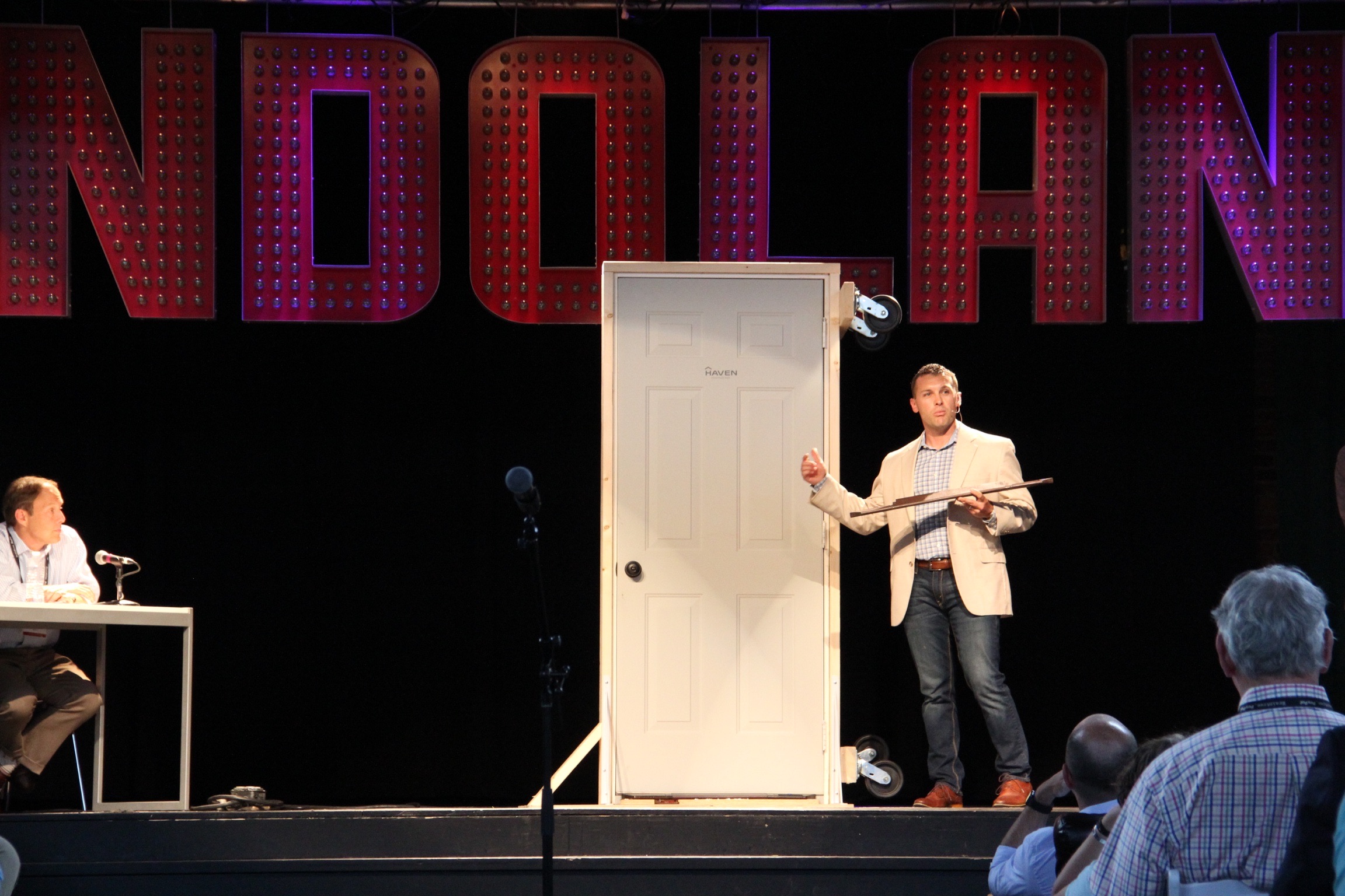 A man in a tan blazer stands on a stage next to a white door mounted on a metal frame, presenting to an audience.